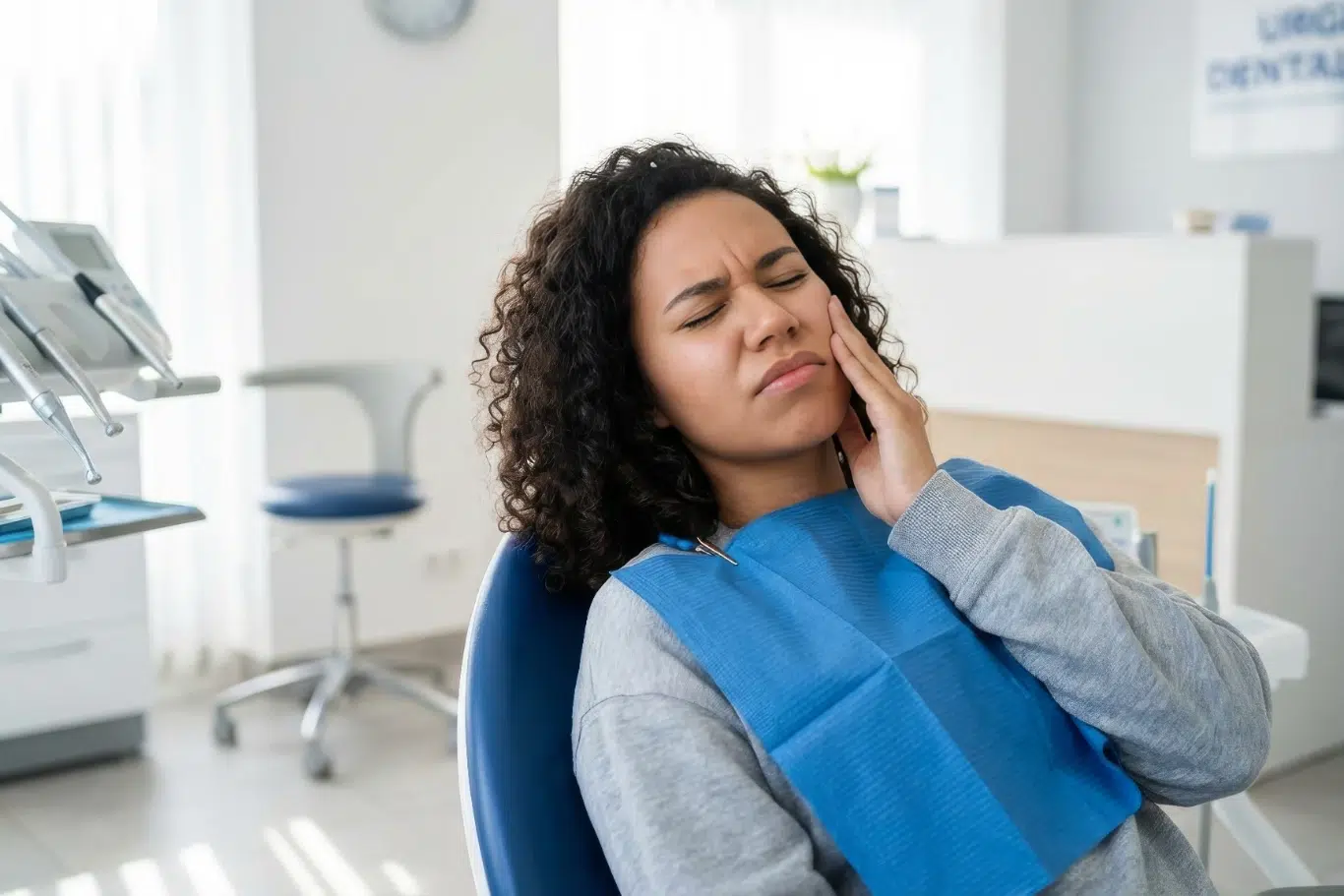Female Patient Experiencing Acute Toothache in Dental Clinic A young woman with curly hair sitting in a dental chair wearing a blue bib grimacing in pain and holding her cheek